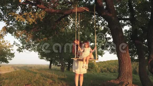 快乐的妈妈把一个快乐的女儿卷在橡树上的公园里的秋千上幸福家庭的概念单身母亲视频的预览图