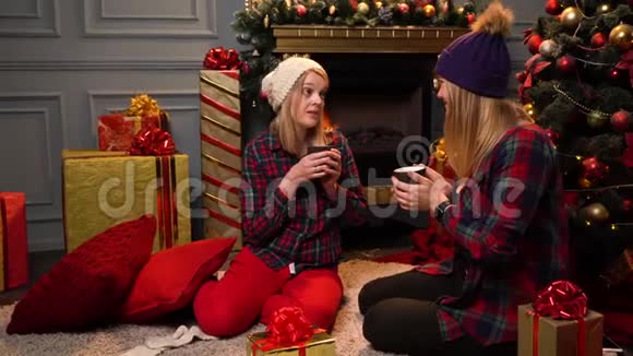 两个穿着红色格子衬衫的女孩坐在圣诞树下戴着温暖的白色和紫色的帽子认真地说话视频的预览图
