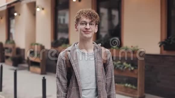 戴着T恤和夹克的眼镜戴着红色卷发站在老城区的年轻微笑男子的肖像视频的预览图