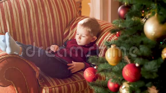 小男孩躺在圣诞树旁边的沙发上用平板电脑和浏览互联网的4K镜头儿童视频的预览图