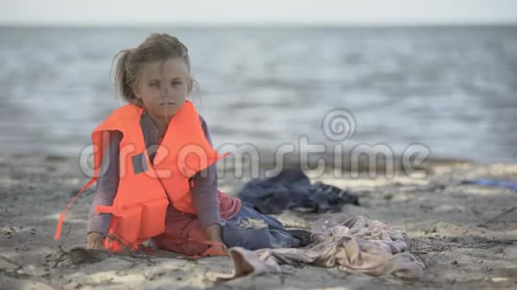 孤独的儿童难民穿着救生衣坐在岸边危险地跨海迁徙视频的预览图