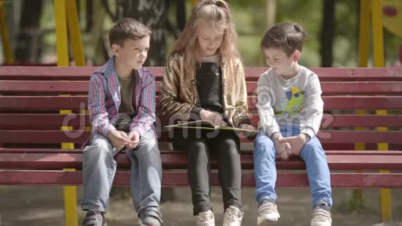 可爱的少女正在户外坐在长凳上给两个弟弟读一本书孩子们在阳光下休闲视频的预览图