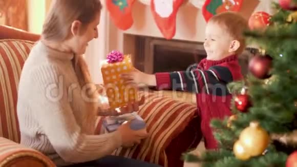 4k视频快乐微笑的小男孩把圣诞礼物盒配丝带送给圣诞树旁的妈妈视频的预览图