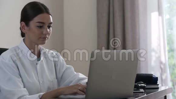 专业成功的女医生坐在办公室的桌子旁在笔记本电脑上打字一个年轻的女孩视频的预览图