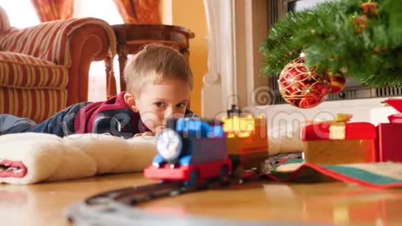 小男孩躺在圣诞树下的地板上看着火车上的玩具火车接收儿童视频的预览图