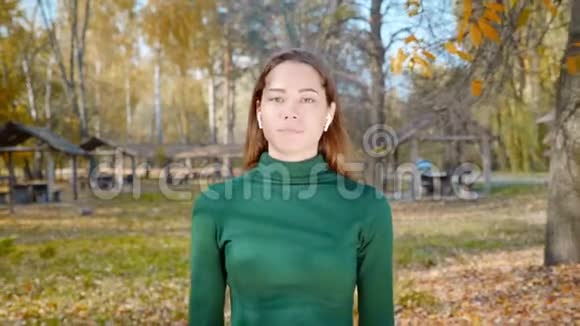 秋天一位年轻漂亮的黑发女人带着无线耳机在公园里散步公园里的树木已经发黄视频的预览图