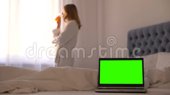 床上的笔记本电脑靠近窗户的女士喝果汁放松在线旅行预订视频的预览图