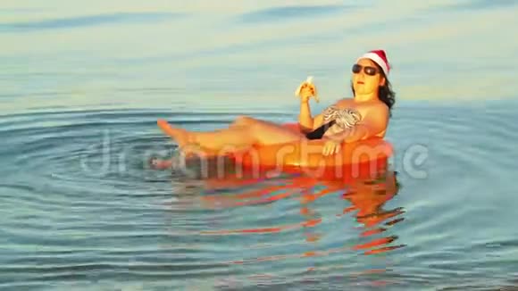 一个身穿泳装戴墨镜的黑发女人在游泳圈里戴着圣诞帽的圣诞老人在圣诞节吃香蕉视频的预览图