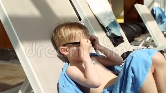 一个金发的小男孩戴着太阳镜在阳光休息室里晒太阳视频的预览图