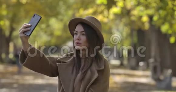 年轻时尚的高加索女人戴着棕色帽子在公园的智能手机上自拍美丽的欧洲女孩视频的预览图