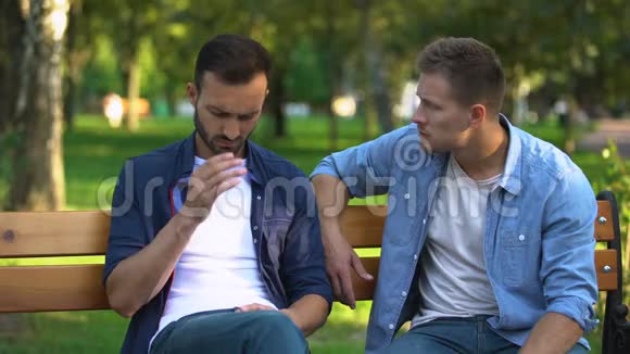 男人安慰悲伤的男性朋友坐在公园的长凳上生活问题支持视频的预览图