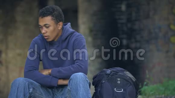 受伤的男孩咳嗽坐在背包附近的街道上从家里跑出来视频的预览图