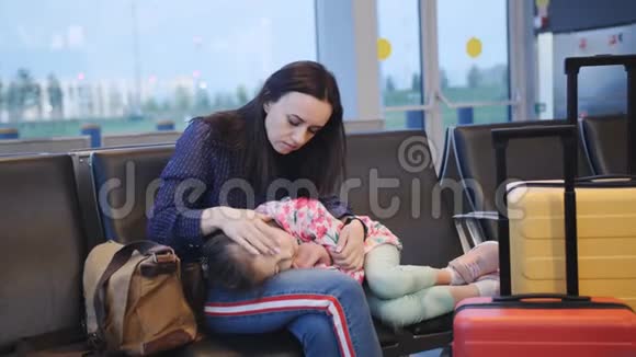 一位母亲抱着她的小女儿在机场候机楼的椅子上睡觉等着她的下一个视频的预览图