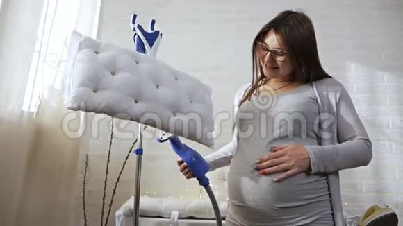 美丽的年轻孕妇白色背景的灰色西装用蒸汽系统在家里熨烫枕头视频的预览图