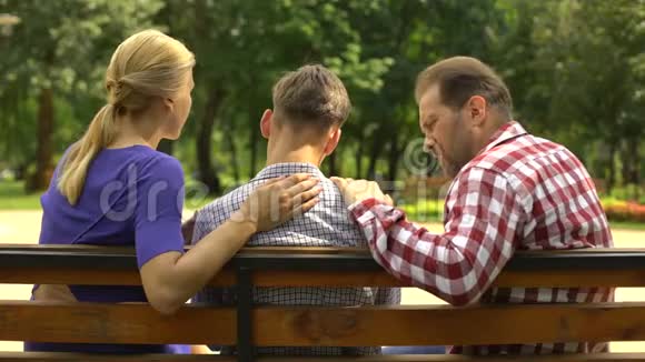 关爱的母亲和父亲支持悲伤的少年儿子坐在公园的长凳上危机视频的预览图