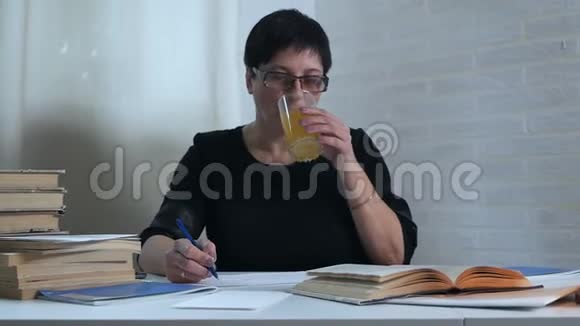 一个穿着黑色衣服的女人坐在白色背景的桌子旁在纸上做笔记喝一杯水视频的预览图