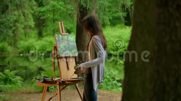 一位年轻的艺术家女士在夏林中画一幅风景画的后中景视频的预览图