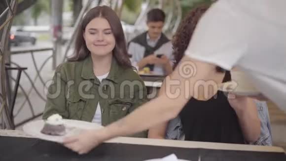 两个可爱的女孩坐在户外咖啡馆里吃蛋糕服务员端着盘子和甜点摆在桌上视频的预览图