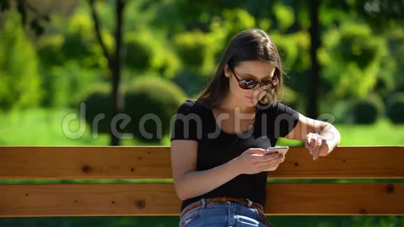 戴墨镜的女孩在阳光公园里坐在长凳上滚动智能手机放松视频的预览图