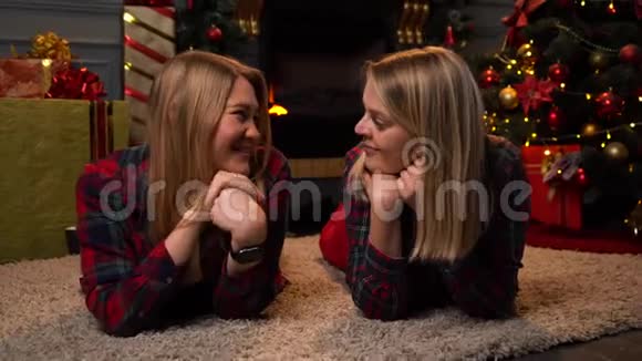 两个穿着红色格子衬衫的金发美女躺在靠近新年树或圣诞树的地毯上互相看着对方视频的预览图