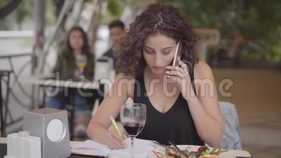 可爱的卷发女孩写下坐在咖啡馆里的桌子上用手机聊天的信息忙碌的自由职业者视频的预览图