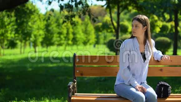 病人女孩看手表等朋友坐在公园的长凳上视频的预览图