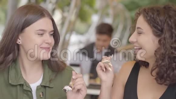 两个女朋友坐在户外的咖啡馆里互相吃蛋糕的肖像年轻女孩一起享受视频的预览图