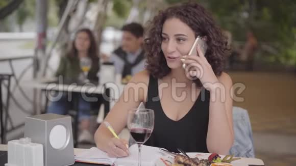 漂亮的卷发女孩在咖啡馆里坐在桌子旁用手机聊天写下信息忙碌的自由职业者视频的预览图
