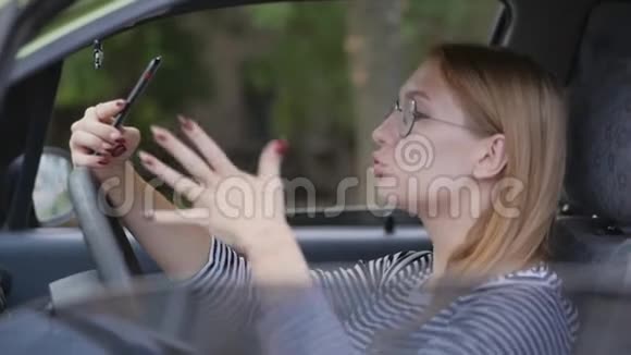 一个年轻的女孩坐在汽车的车轮后面用智能手机里的摄像机进行情感交谈用手势表示视频的预览图