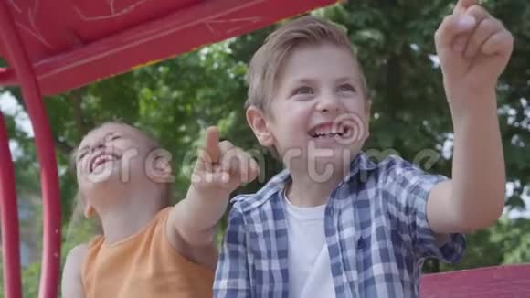 可爱的男孩和女孩指着一边手指坐在公园里的秋千上开心地笑着一对夫妇视频的预览图