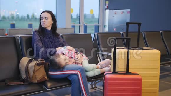 一位母亲抱着她的小女儿在机场候机楼的椅子上睡觉等着她的下一个视频的预览图