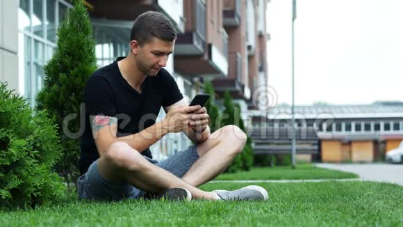 有魅力的男人坐在酒店附近的草地上和朋友聊天时使用手机视频的预览图