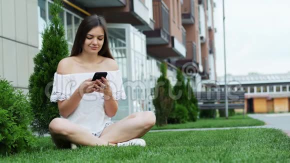 可爱的女孩坐在酒店附近的草地上和朋友聊天时使用手机视频的预览图