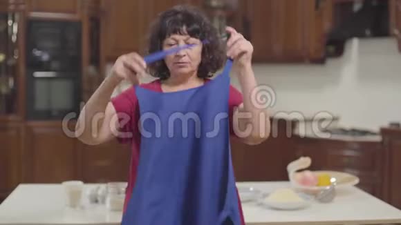 现代厨房里穿蓝色围裙的白种人成熟女人的肖像快乐的高级退休人员视频的预览图