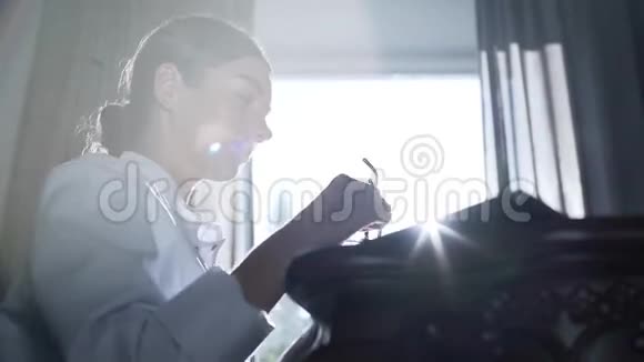 职业女医生戴着眼镜在笔记本电脑上打字坐在靠窗的桌子旁视频的预览图