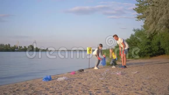 志愿者的家人年轻的父亲和母亲带着小女儿在肮脏的海滩上的垃圾袋里收集垃圾视频的预览图