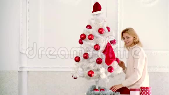 准备圣诞礼物金发女人在白色的圣诞树上拿着红色的圣诞球圣诞装饰品视频的预览图