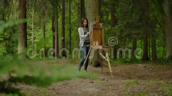一位可爱的艺术家在森林的画架上画一幅风景画视频的预览图