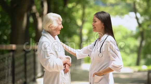 微笑护士与身穿浴袍的老妇人在疗养院公园交谈放松视频的预览图