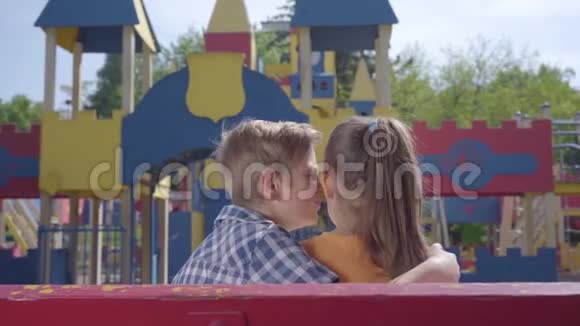 可爱的金发男孩亲吻一个漂亮女孩的脸颊坐在操场前的长凳上一对快乐视频的预览图