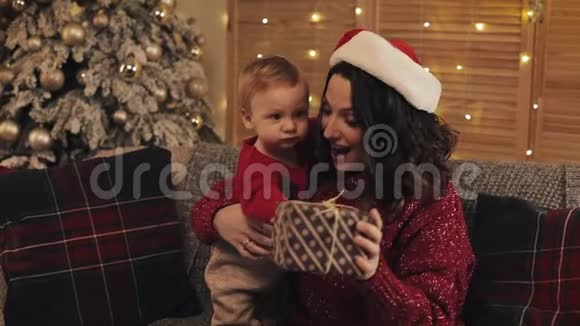 年轻漂亮的妈妈戴着圣诞帽和可爱的小男孩坐在沙发上看着圣诞树旁快乐视频的预览图