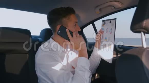年轻的商人与司机一起驾驶汽车讨论他的手机上的统计和工作结果视频的预览图