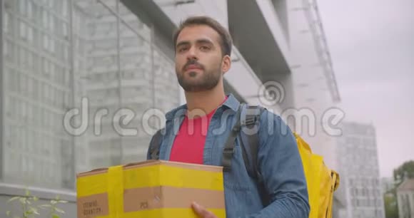 留胡子的白种人送货员的特写肖像背着背包的箱子走在城市的街道上户外视频的预览图