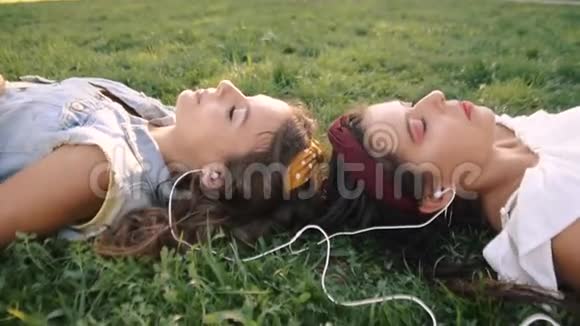 两个女朋友姐妹躺在草地上听着耳机上的音乐两个黑发女人躺在公园的草地上视频的预览图