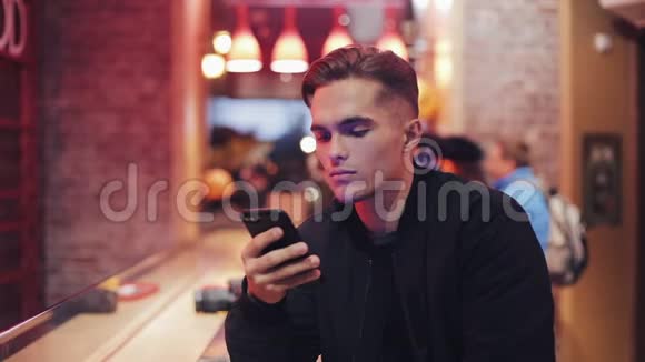 年轻迷人的男人带着美丽的微笑在晚上使用智能手机他坐在靠近霓虹灯的酒吧或餐馆里视频的预览图