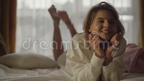 年轻白种人女人躺在柔软的床上和颤抖的腿的肖像穿着白衬衫的微笑女孩在卧室里休息视频的预览图