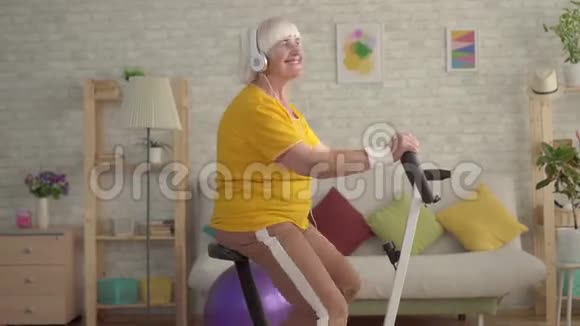 积极积极的祖母耳机听音乐和参与一个固定的自行车关闭视频的预览图