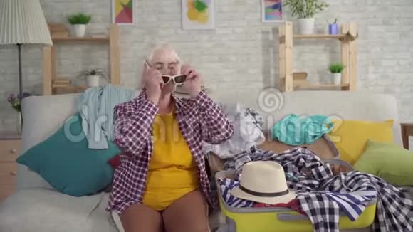 年轻的老年妇女坐在沙发上坐在拆卸的手提箱旁边手里拿着衣服手指朝上视频的预览图