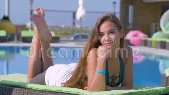在暑假期间长毛快乐的女性在阳光明媚的一天在度假村蓝色游泳池旁的阳光下放松视频的预览图