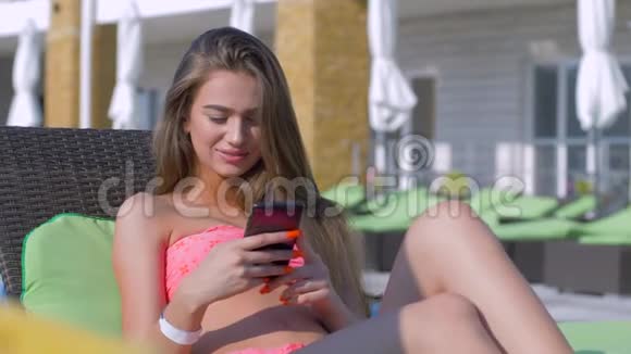 暑假迷人的晒成褐色的年轻女子穿着长发泳衣躺在地上用智能手机聊天视频的预览图